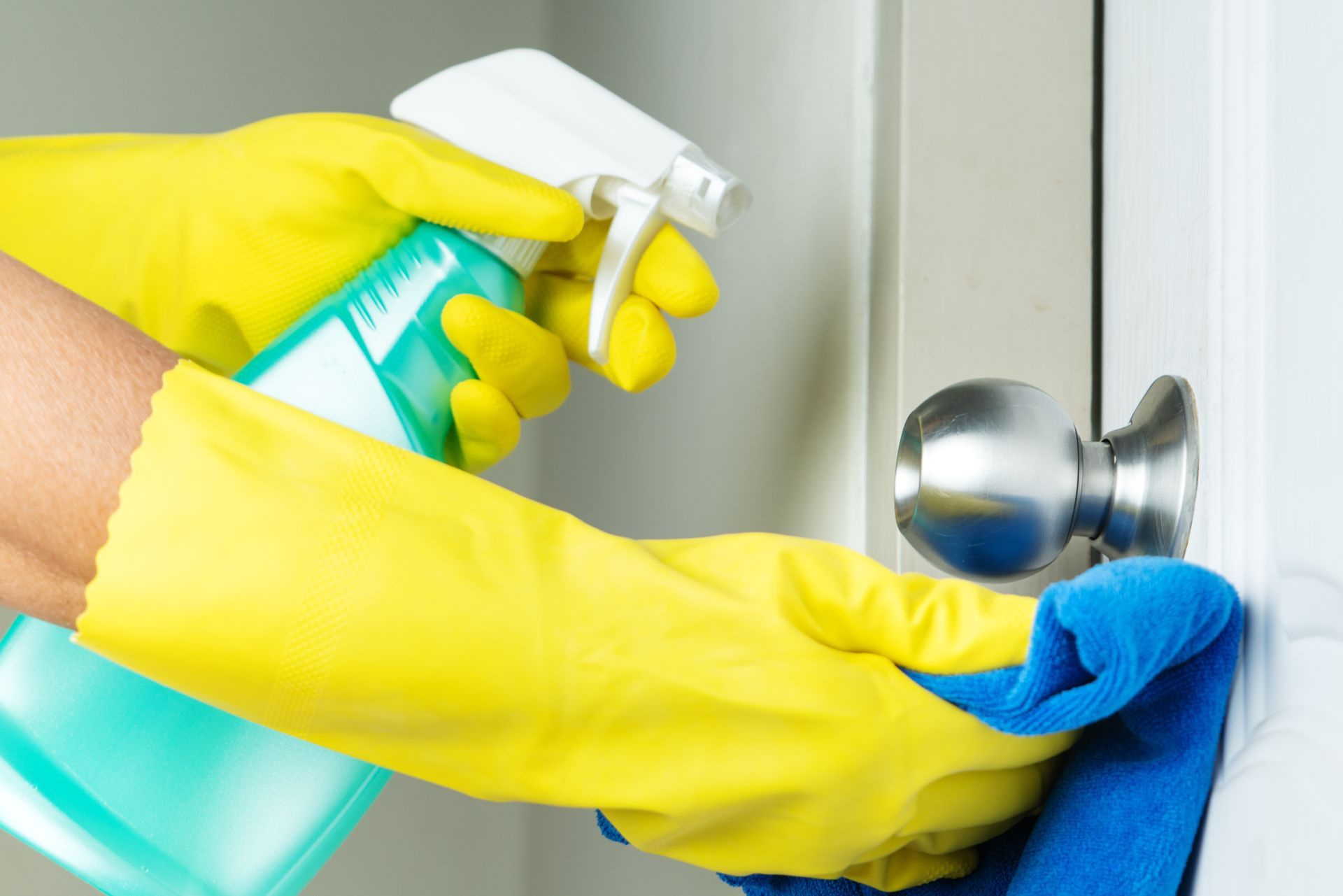 Person in yellow gloves spraying and wiping a doorknob with a blue cloth.