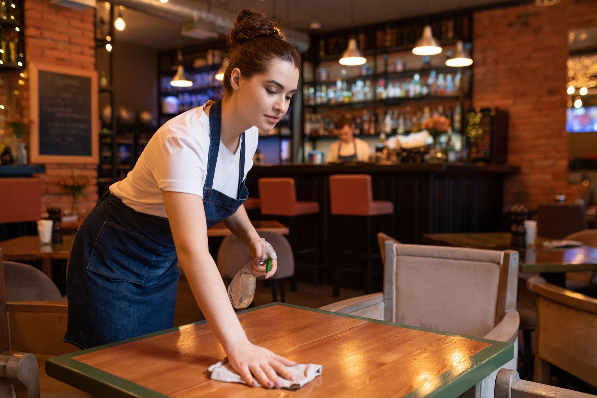 A server in a denim apron cleans a wooden table in a restaurant. A server in a denim apron cleans a wooden table in a restaurant.