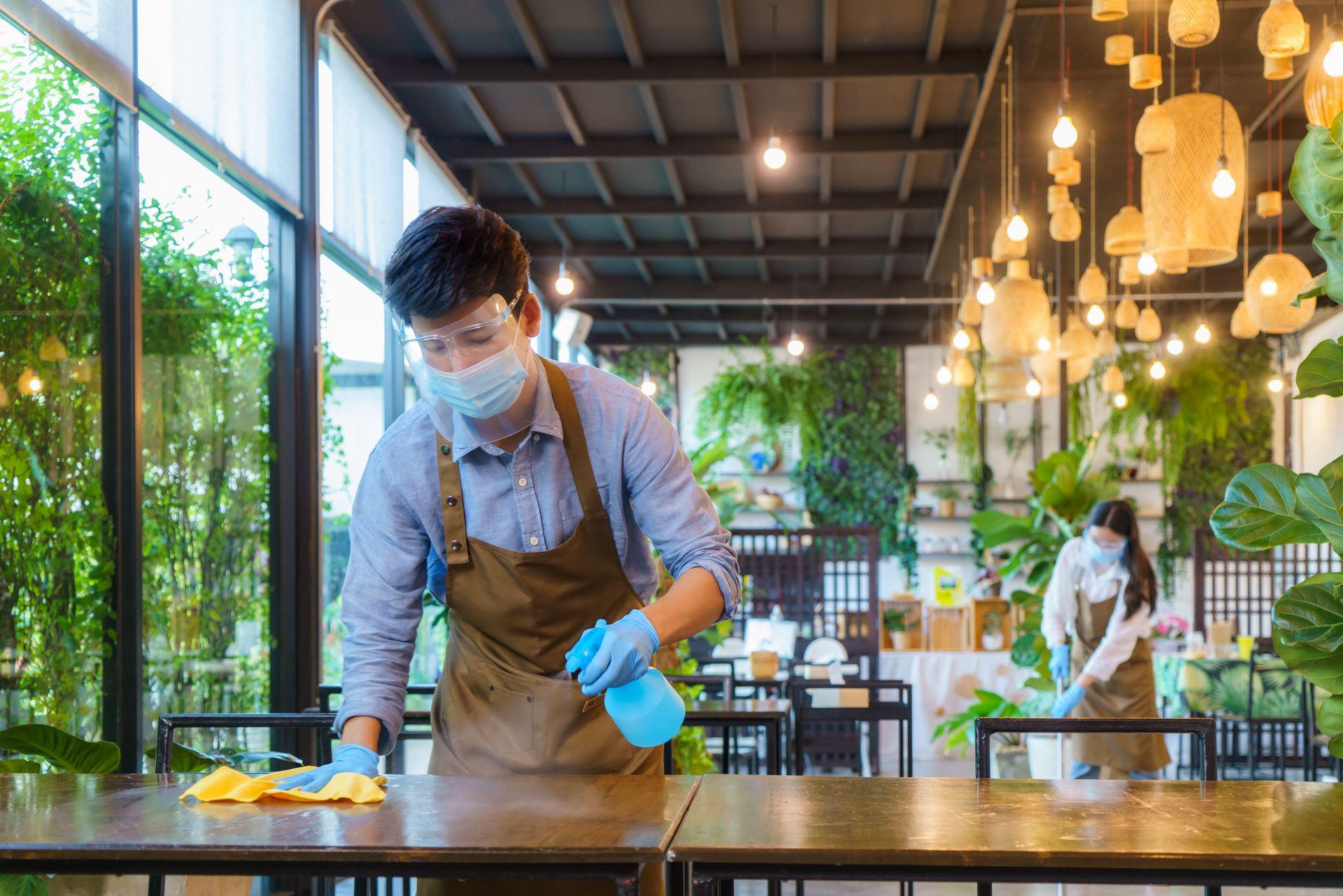 Two masked restaurant workers sanitizing tables with spray bottles and cloths. Two masked restaurant workers sanitizing tables with spray bottles and cloths.