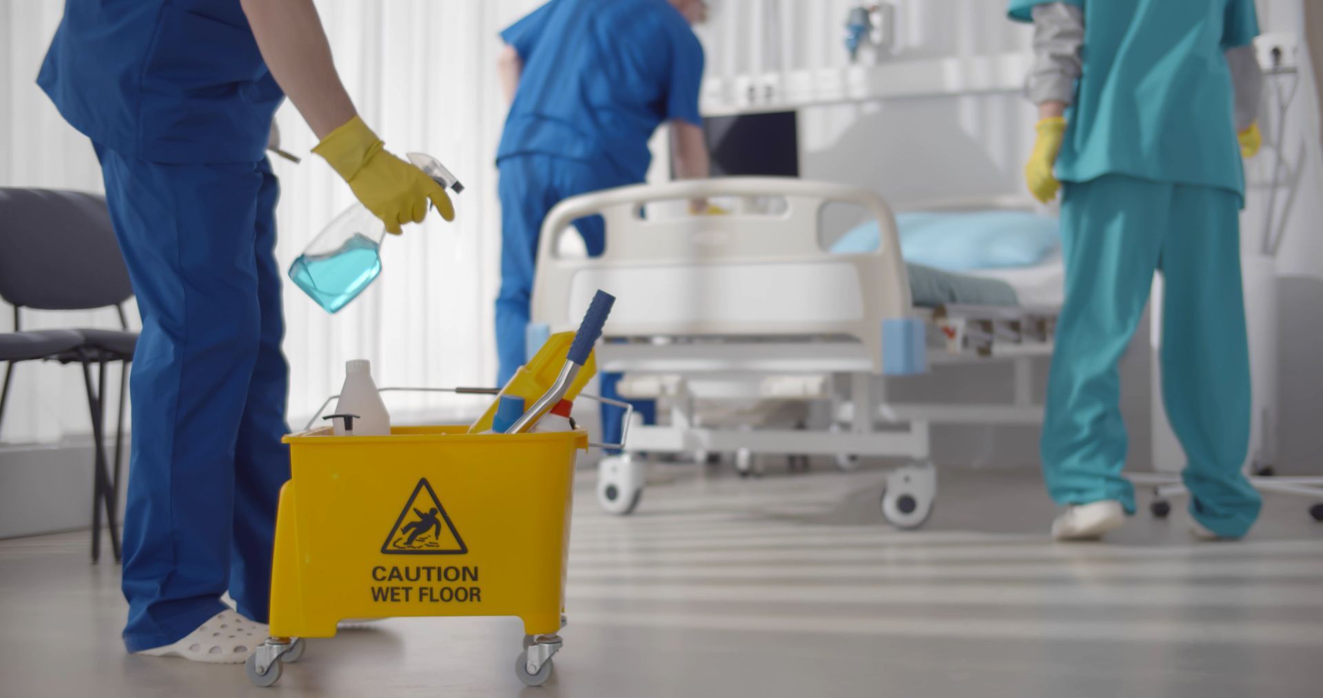 Hospital staff cleaning patient room: person spraying blue liquid into a bucket with cleaning supplies, hospital bed in the background.