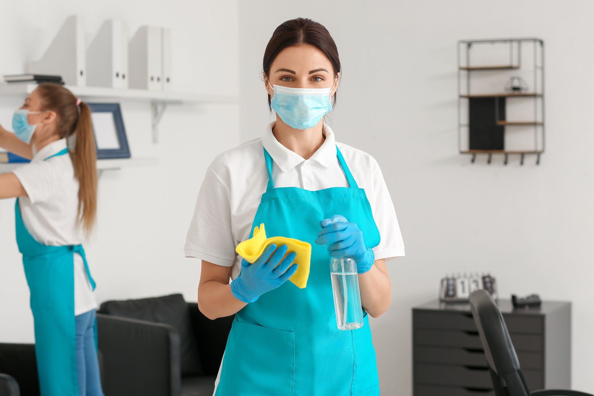 Two cleaning staff wearing aprons and masks, sanitizing an office.