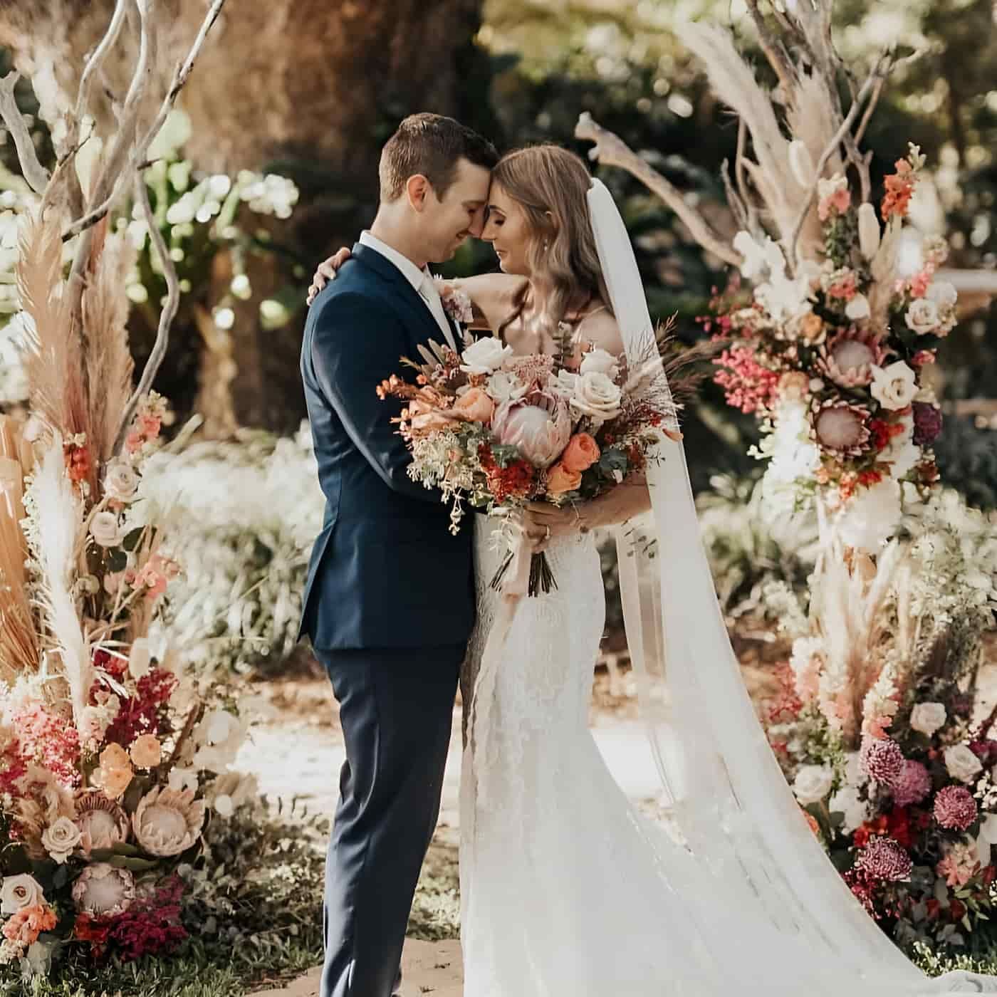 A Bride And Groom Are Posing For A Picture In Front Of A Floral Arch — Lynda Ross Marriage Celebrant In Bowen, QLD