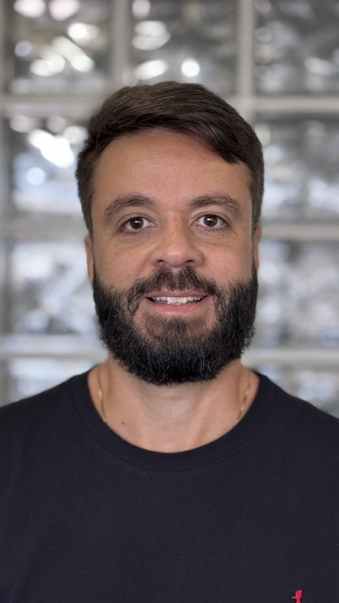 Um homem com barba sorri para a câmera enquanto veste uma camisa preta.