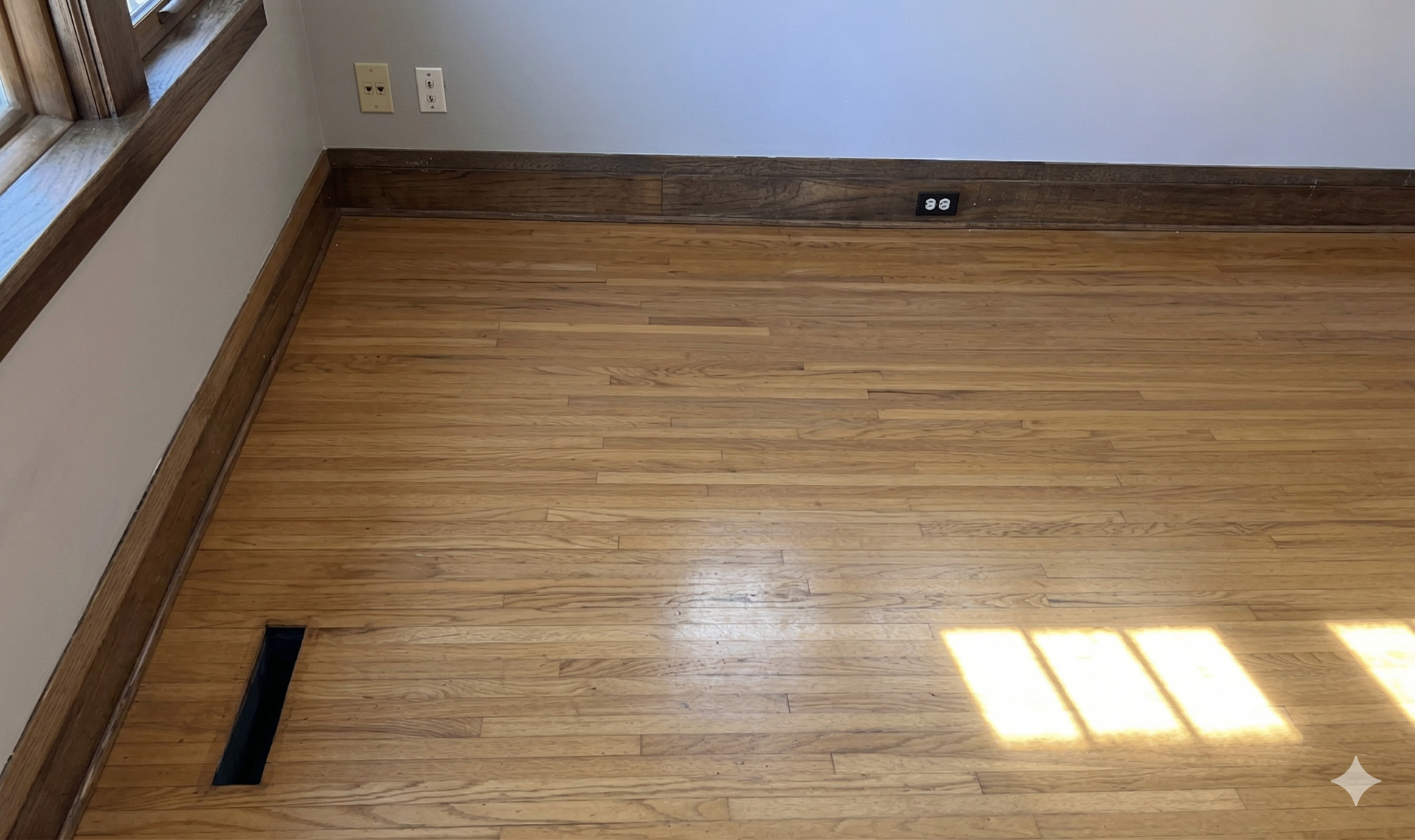 An empty room featuring light-toned wood flooring, dark wood baseboards, a floor vent, and light-colored walls.