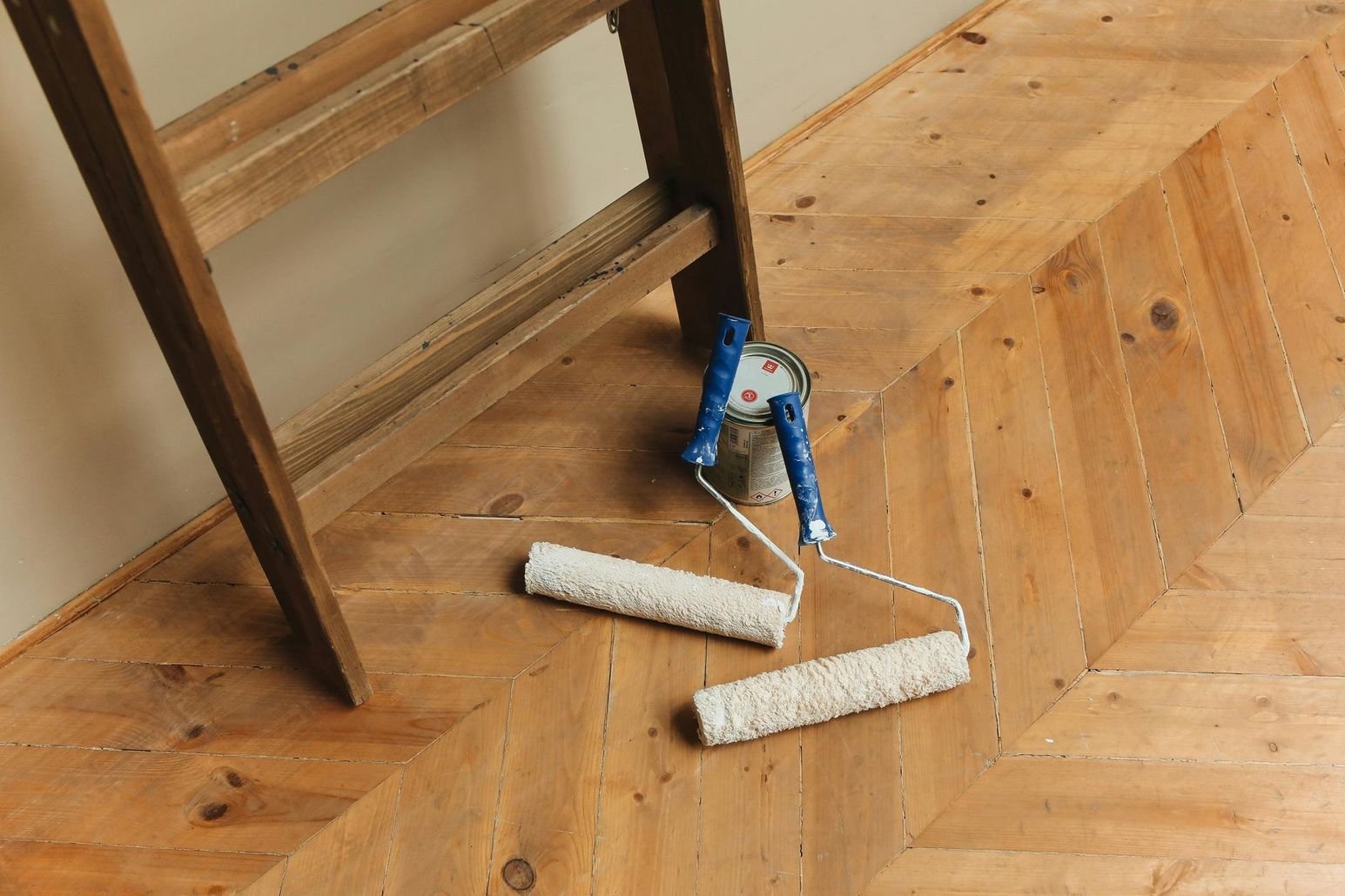 Two paint rollers with blue handles sit on a herringbone-patterned wooden floor near a wooden ladder.