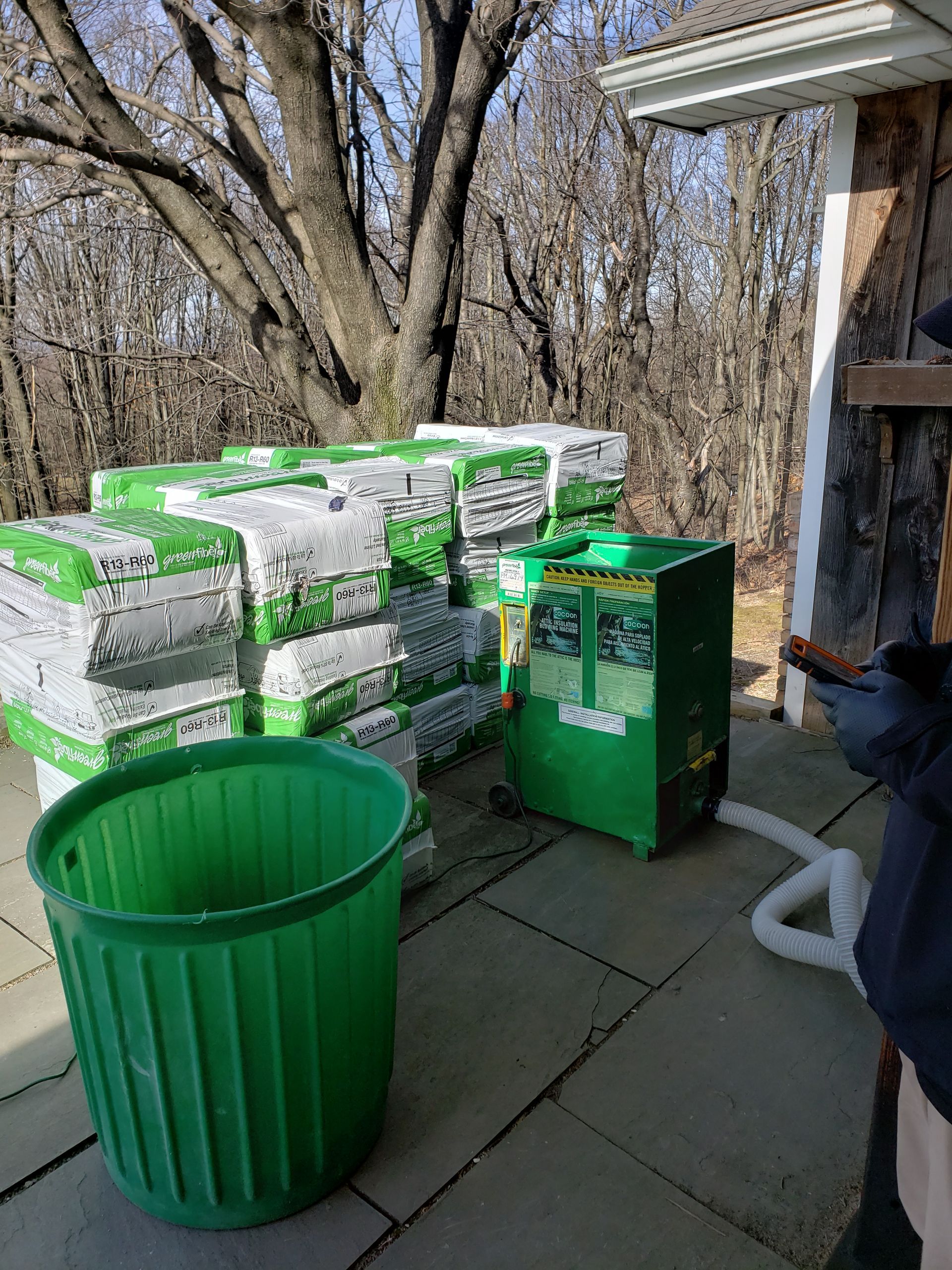 A green trash can is sitting on a patio next to a stack of bags of soil.