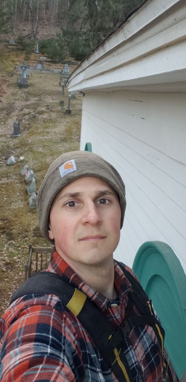 A man wearing a plaid shirt and a beanie is taking a selfie in front of a house.
