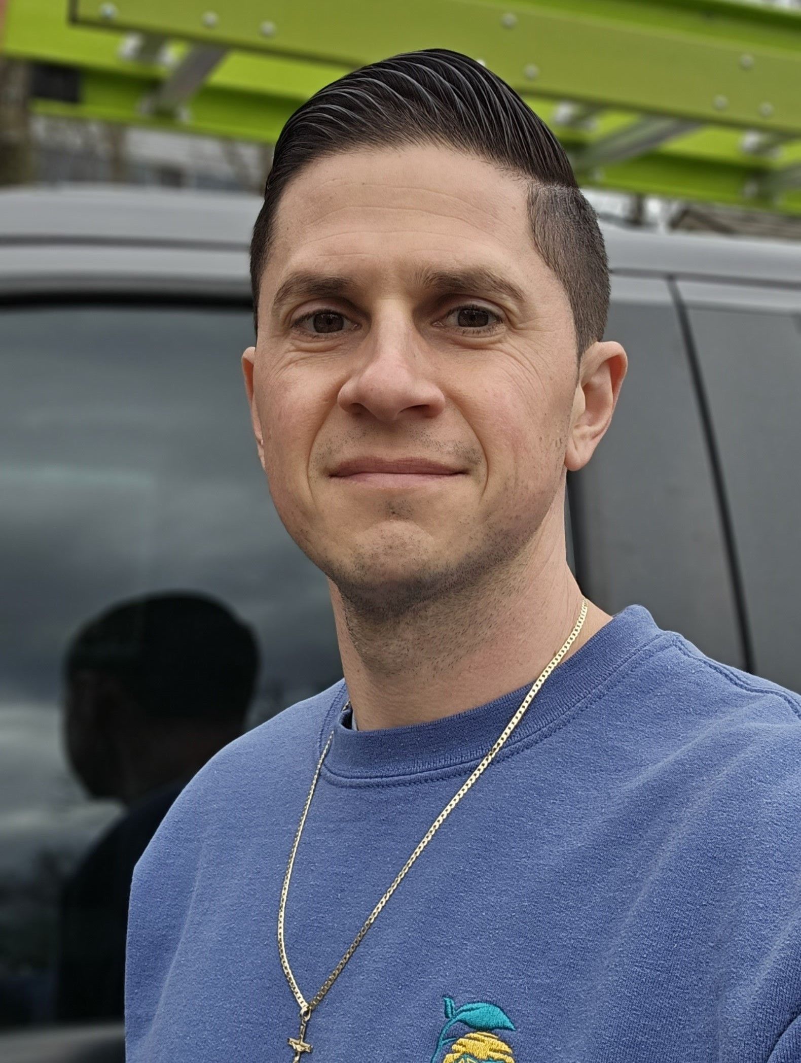 A man wearing a blue shirt and a gold chain is standing in front of a van.