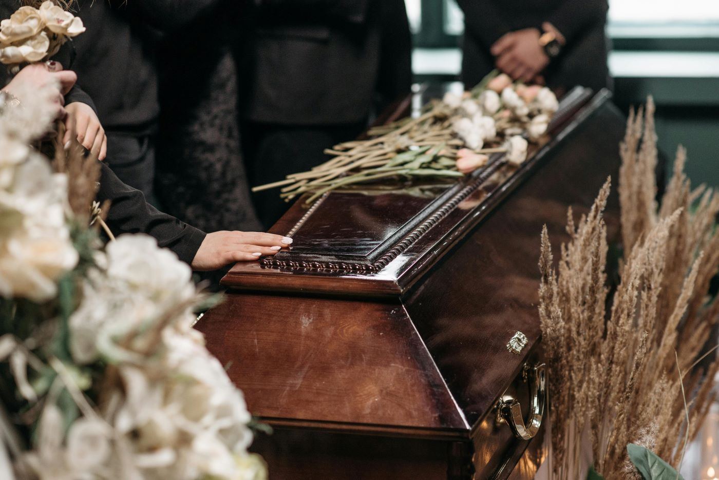 Wooden casket with flowers; a person's hand rests on it during a funeral.