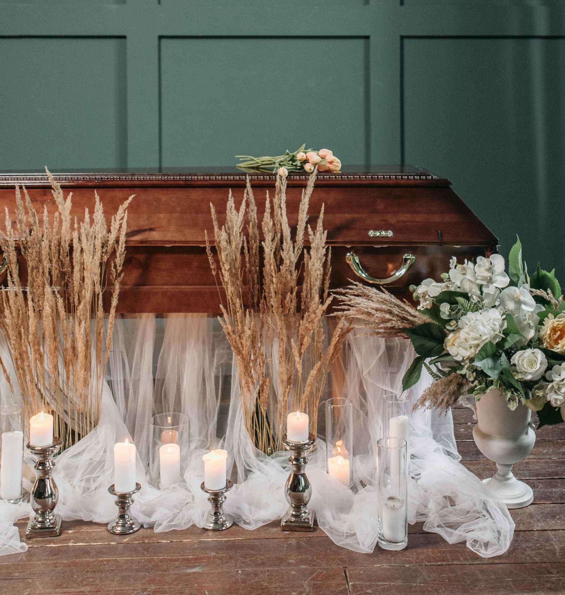 A closed brown casket adorned with flowers, candles, and sheer fabric against a green wall.