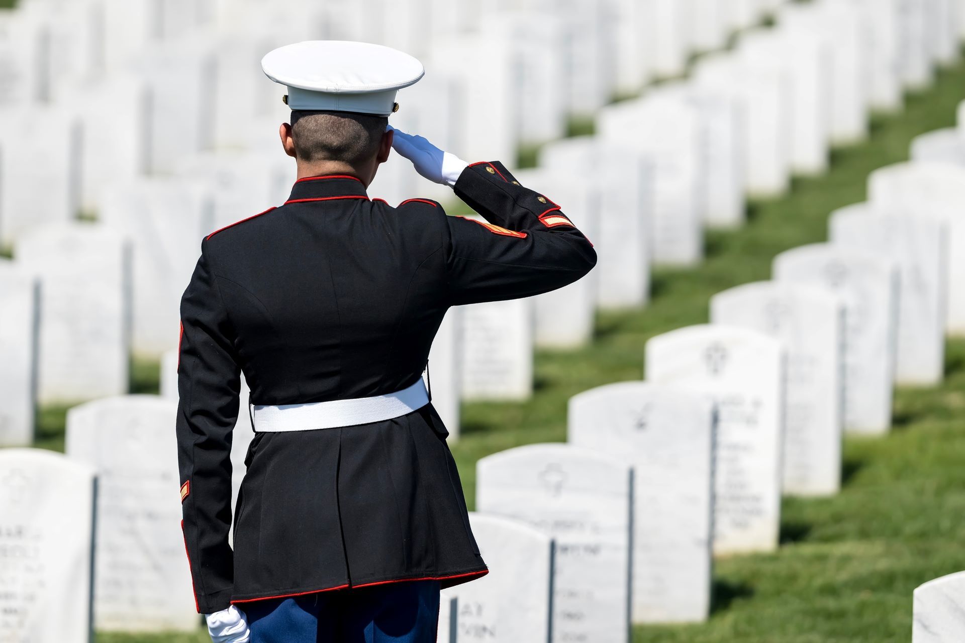 Marine in dress uniform salutes at a cemetery, rows of white headstones in the background.