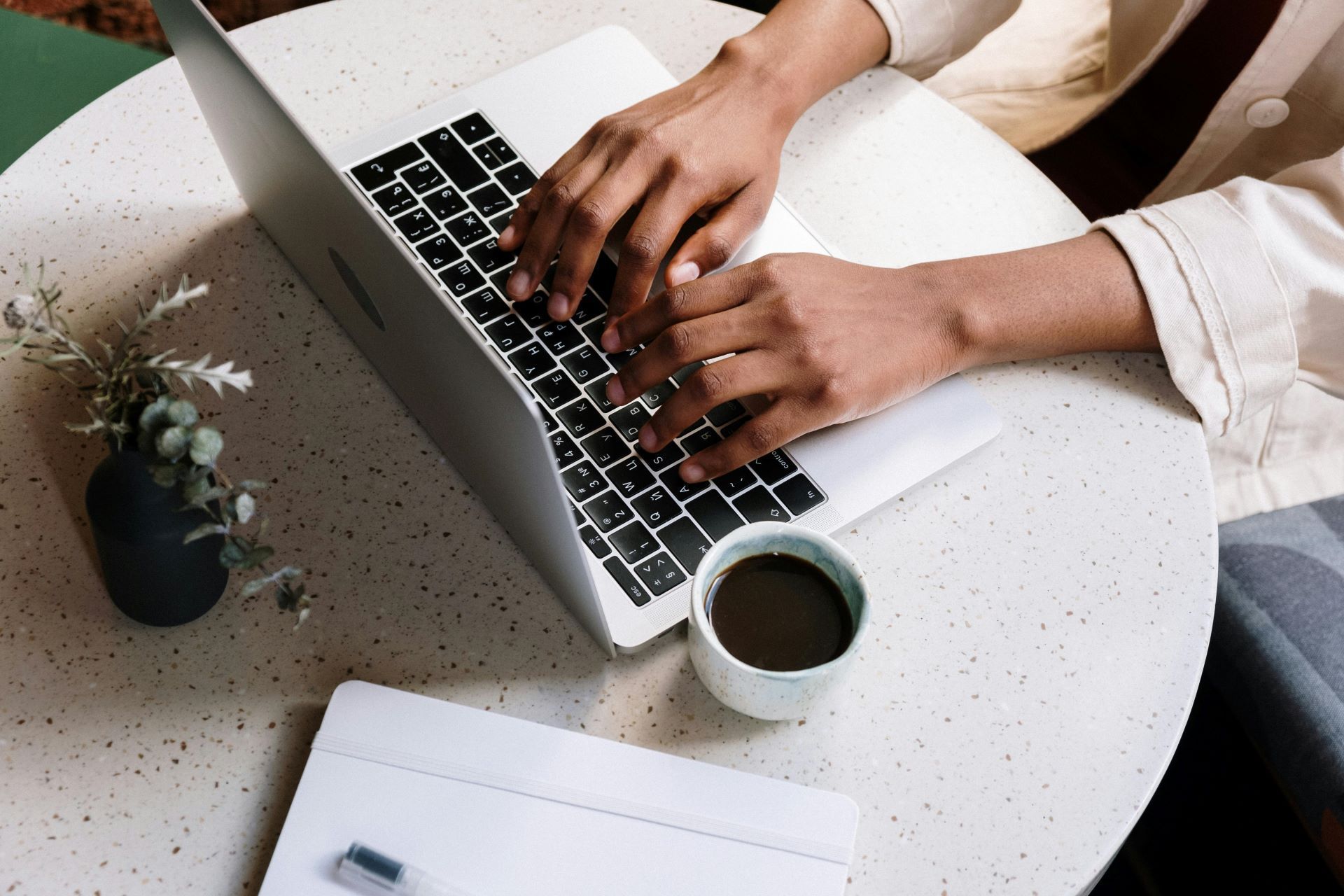 Person typing on a laptop at a table with a coffee cup and small plant.