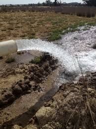 Water gushes from a white pipe, flowing across brown earth in a field.