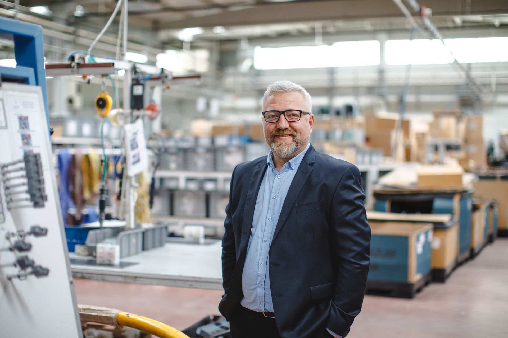 A man in a suit and glasses is standing in a factory with his hands in his pockets.
