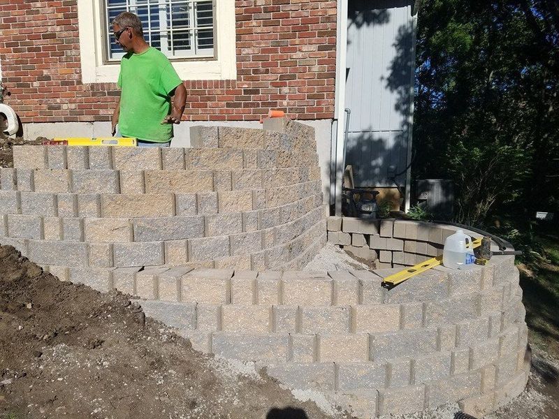 A person in a green shirt building a curved stone retaining wall against a brick house exterior.