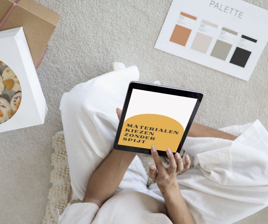 Een persoon die een tablet met tekst vasthoudt, naast een kleurenpalet en een doos gebak, ligt op een kleed.
