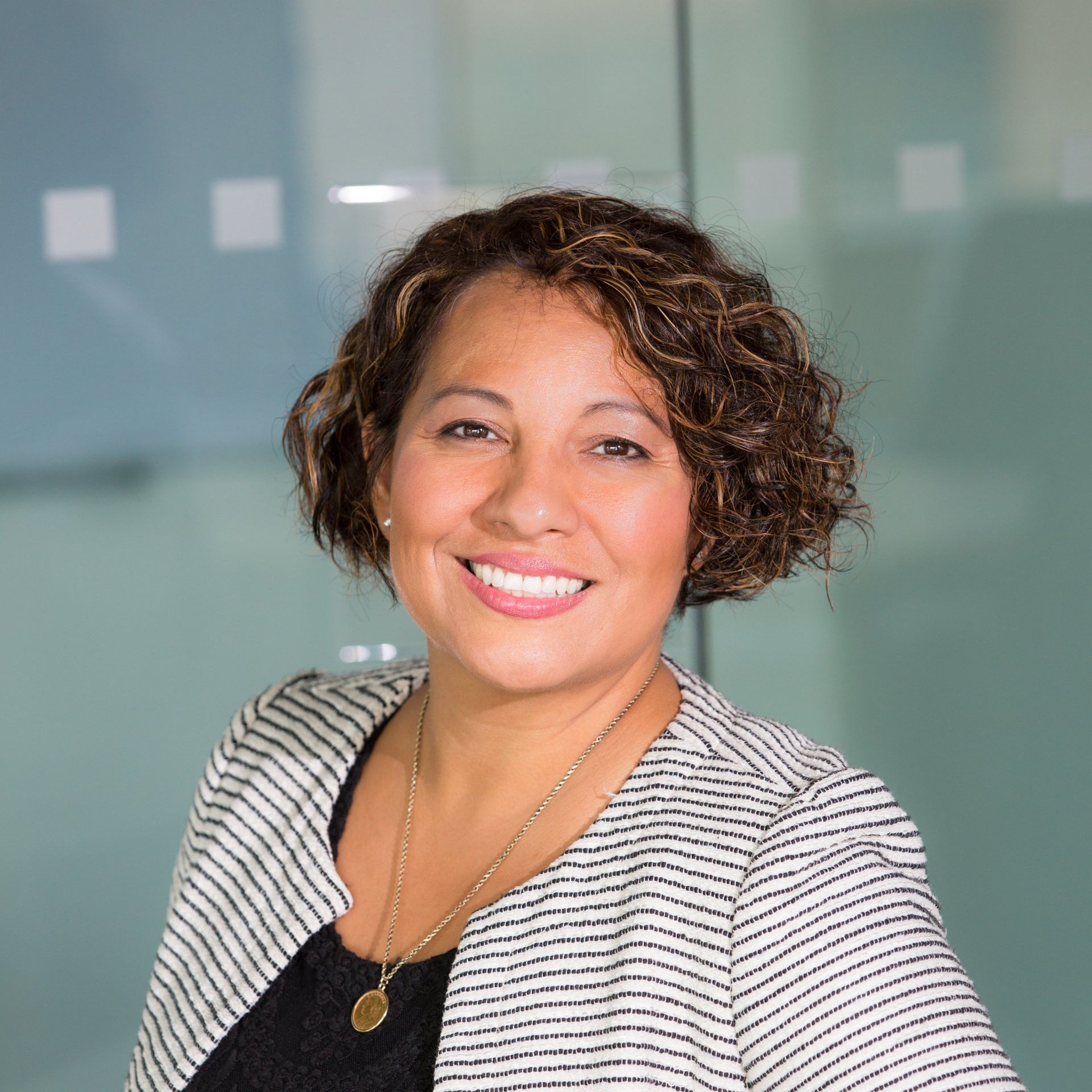 A smiling person with curly dark hair wearing a black top and a black-and-white striped blazer in a bright office.