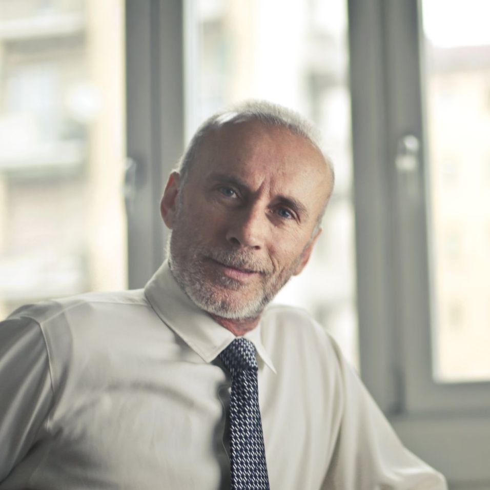A person with a beard, wearing a white shirt and patterned tie, looks toward the camera against a window background.