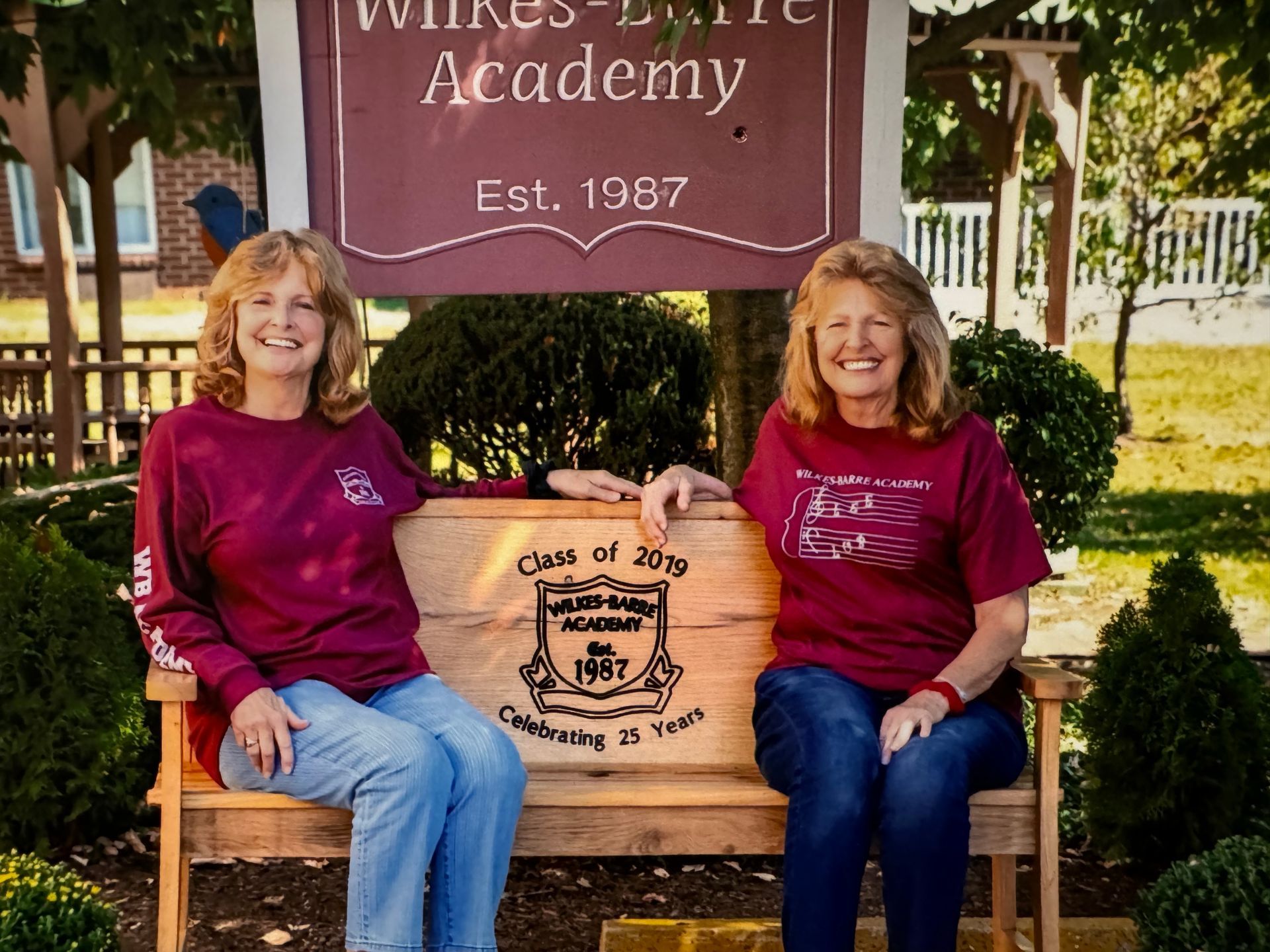 Two women in maroon shirts smiling, sitting on a bench in front of Wilkes-Barre Academy sign.