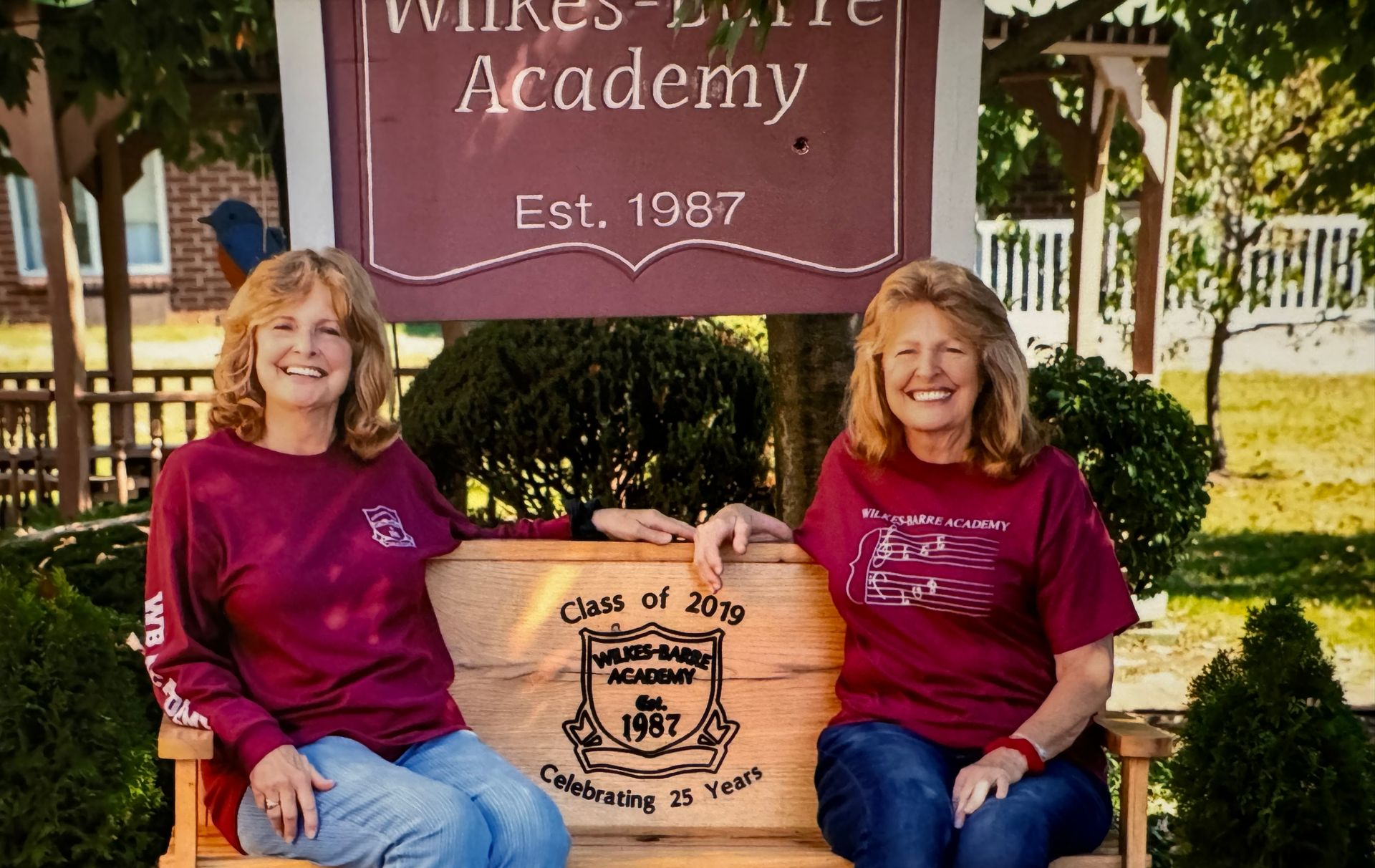 Two women in maroon shirts smiling, sitting on a bench in front of Wilkes-Barre Academy sign.