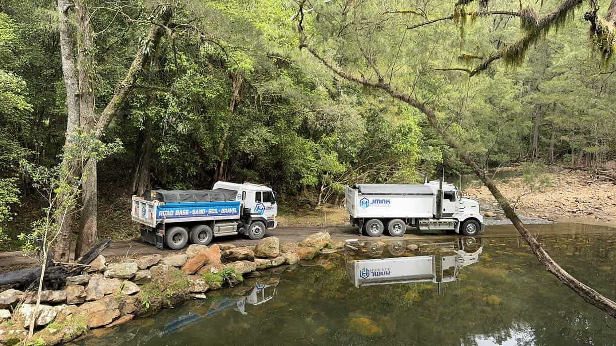 Two Dump Trucks Are Parked Next To A Body Of Water — JMAK Haulage & Landscapes Supplies In Falls Creek, NSW