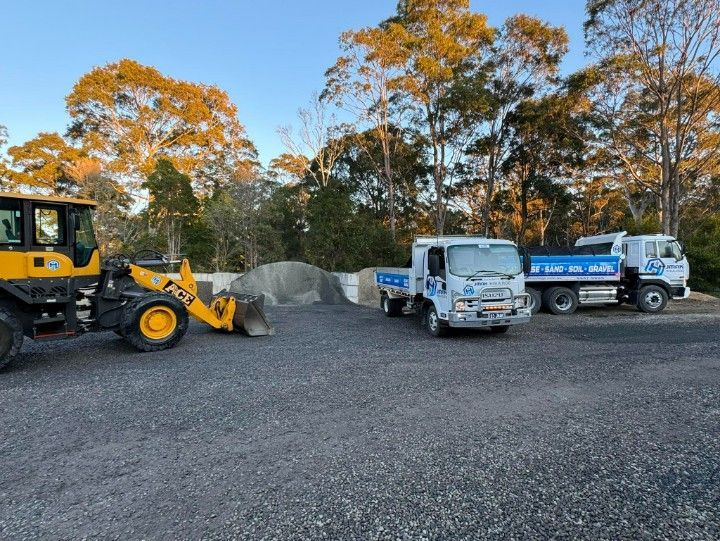 A Yellow Front-end Loader and Two White Trucks Parked on a Gravel Surface — JMAK Haulage & Landscapes Supplies In Ulladulla, NSW