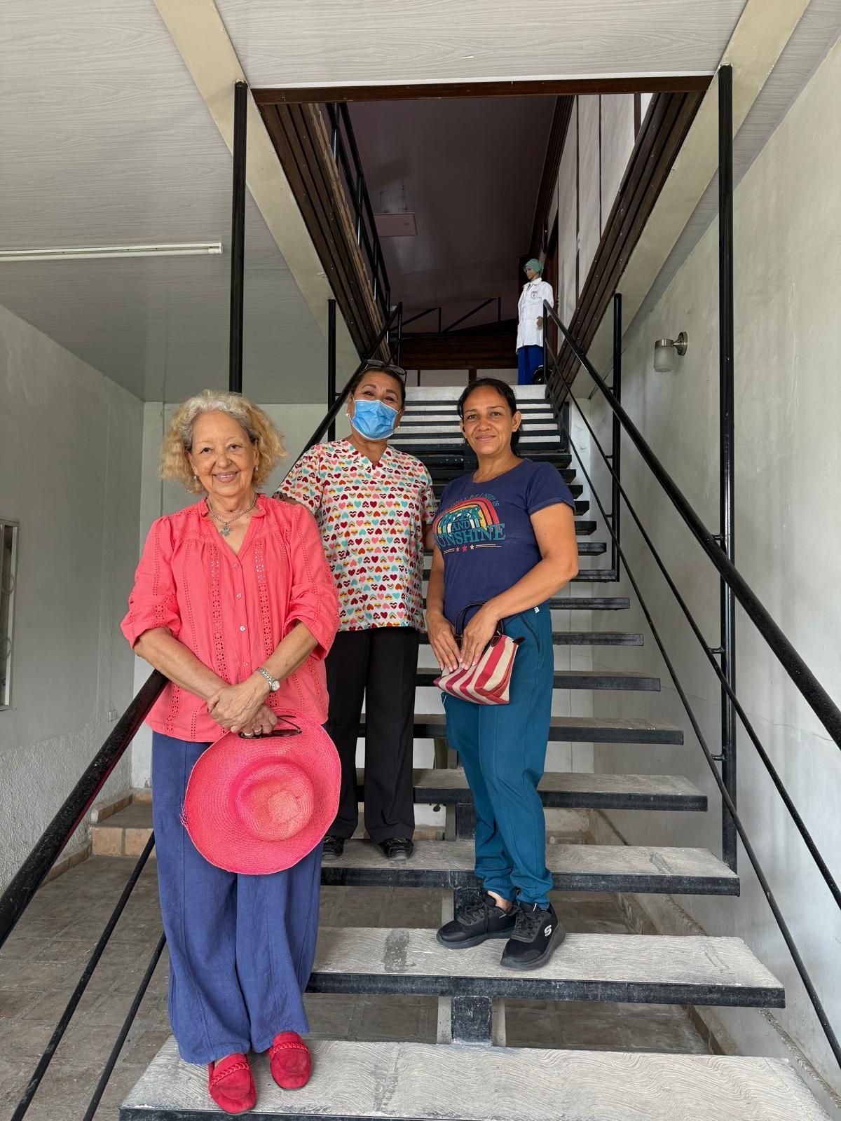 Three women smiling on stairwell, one holding a pink hat.