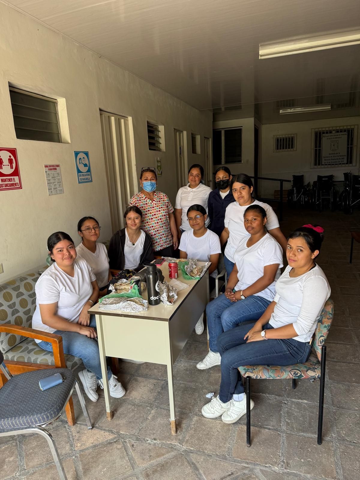 Group of people, mostly women, in white shirts and jeans pose near a table outdoors.