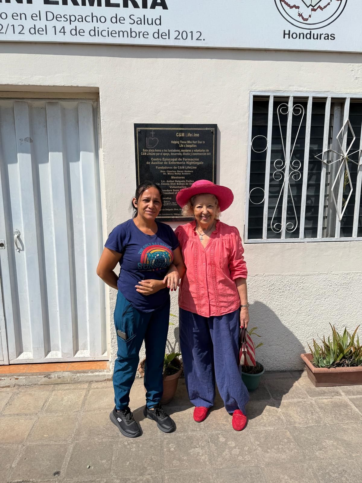 Two women pose in front of a building in Honduras. One wears a blue shirt and pants, the other a pink shirt, blue pants and a hat.