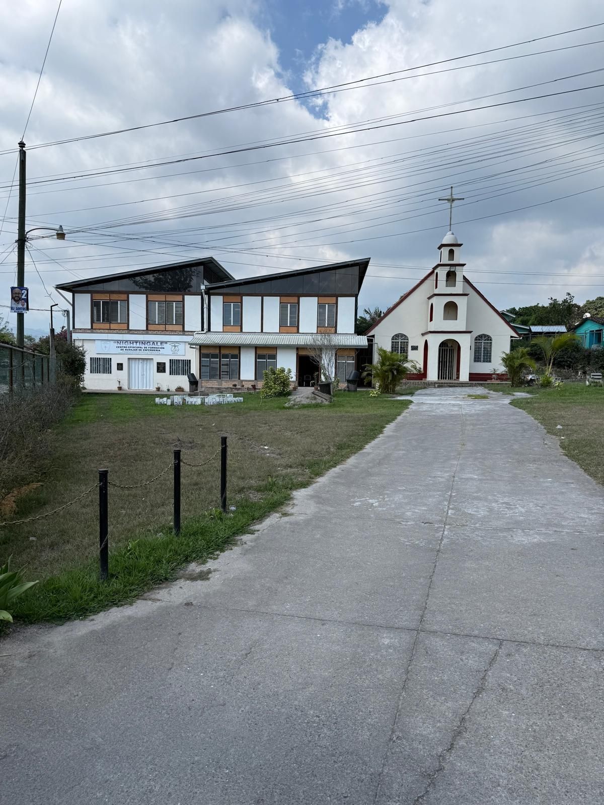 Church and adjacent building on a grassy lot under a cloudy sky. A paved path leads to the church.