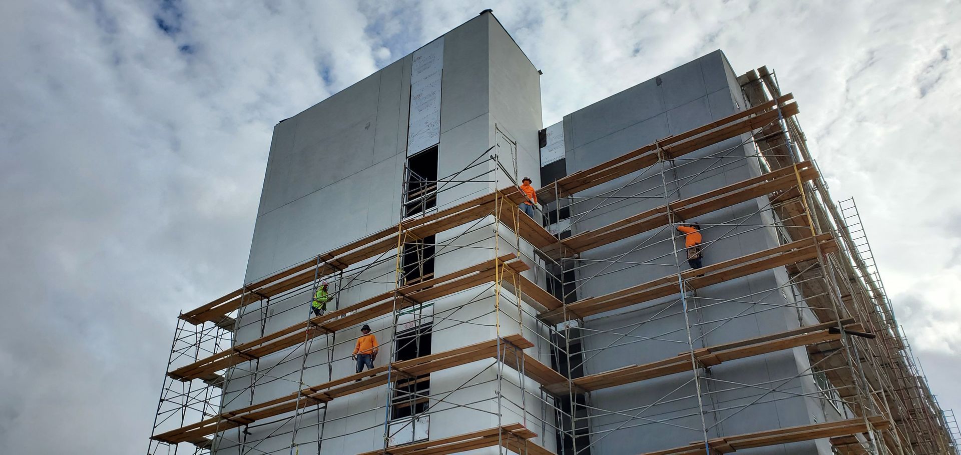 Construction workers on scaffolding against a modern building under a cloudy sky.