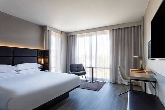 Hotel room with bed, chair, desk, and large window with light-colored curtains.