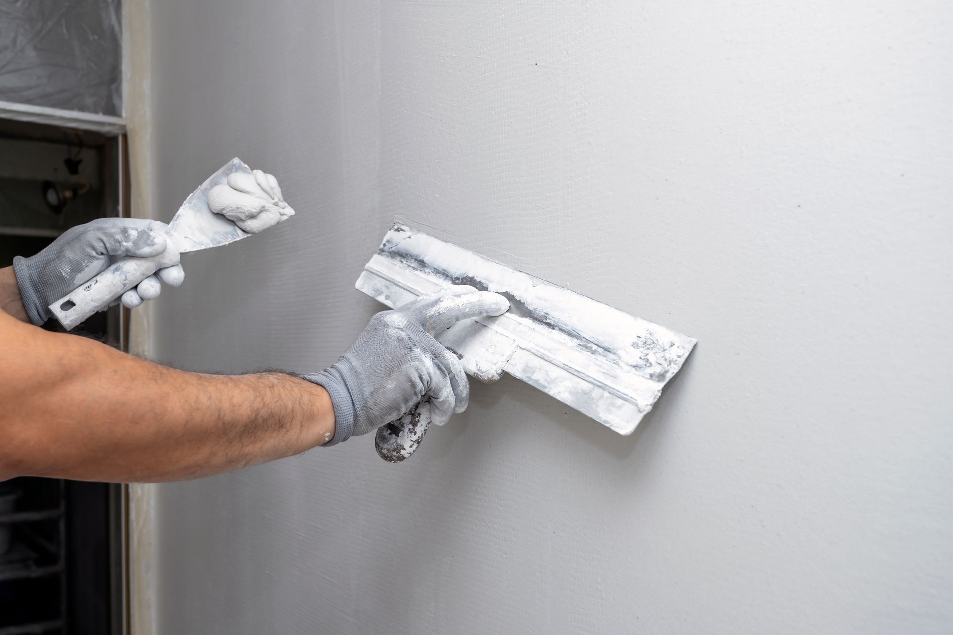 Person wearing gloves smoothing wall with joint compound and two putty knives.