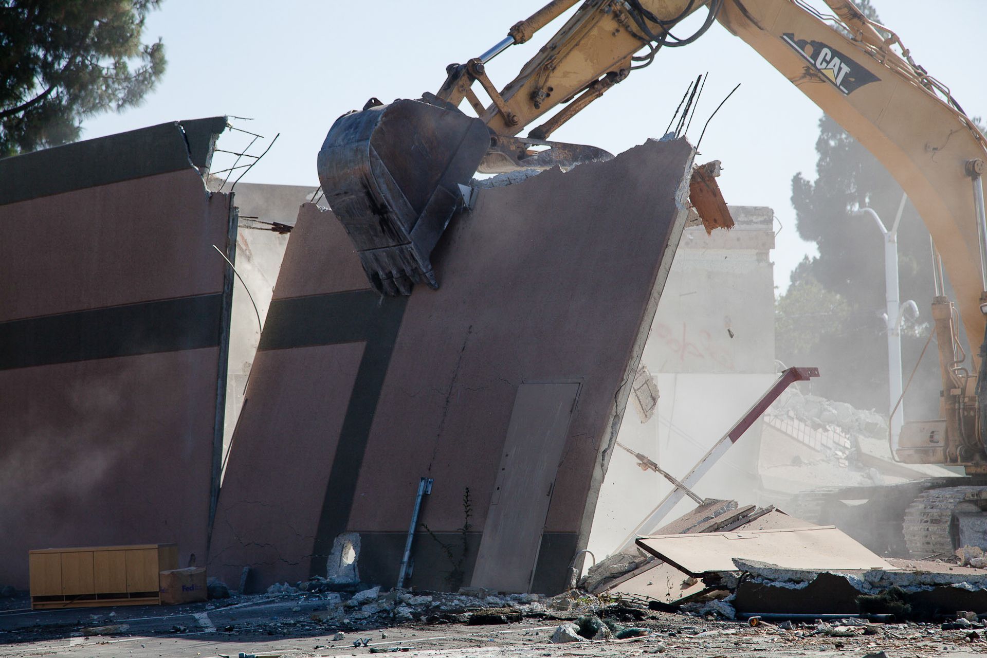 An excavator demolishes a concrete wall, kicking up dust.