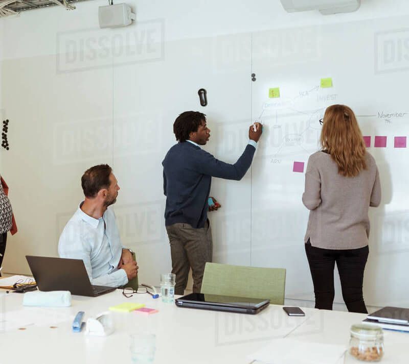 Person in a dark shirt facing a whiteboard with red diagrams, holding papers, indoors.