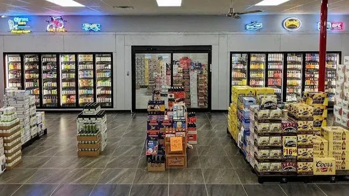 Inside a liquor store: cold beverage refrigerators flank a central aisle; various products displayed on pallets and shelves.