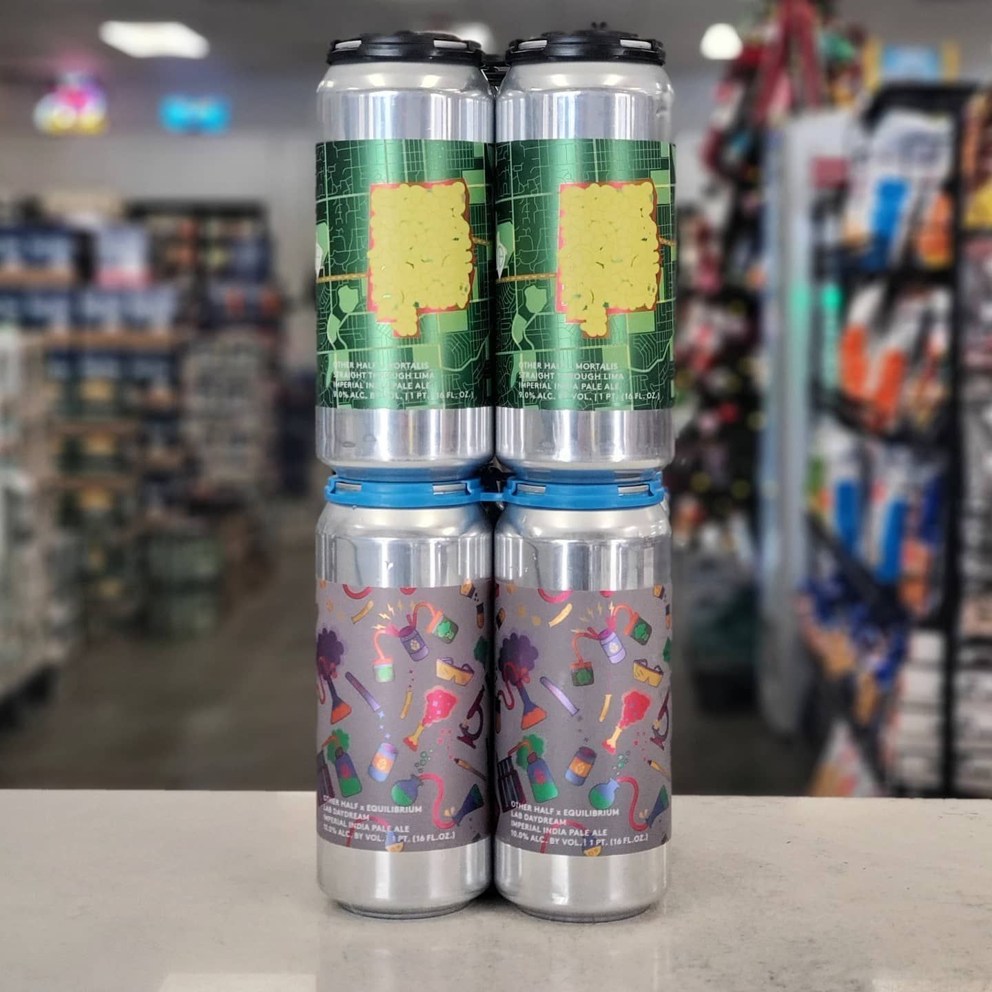 Four beer cans stacked. Green and gray label designs, in a store setting.