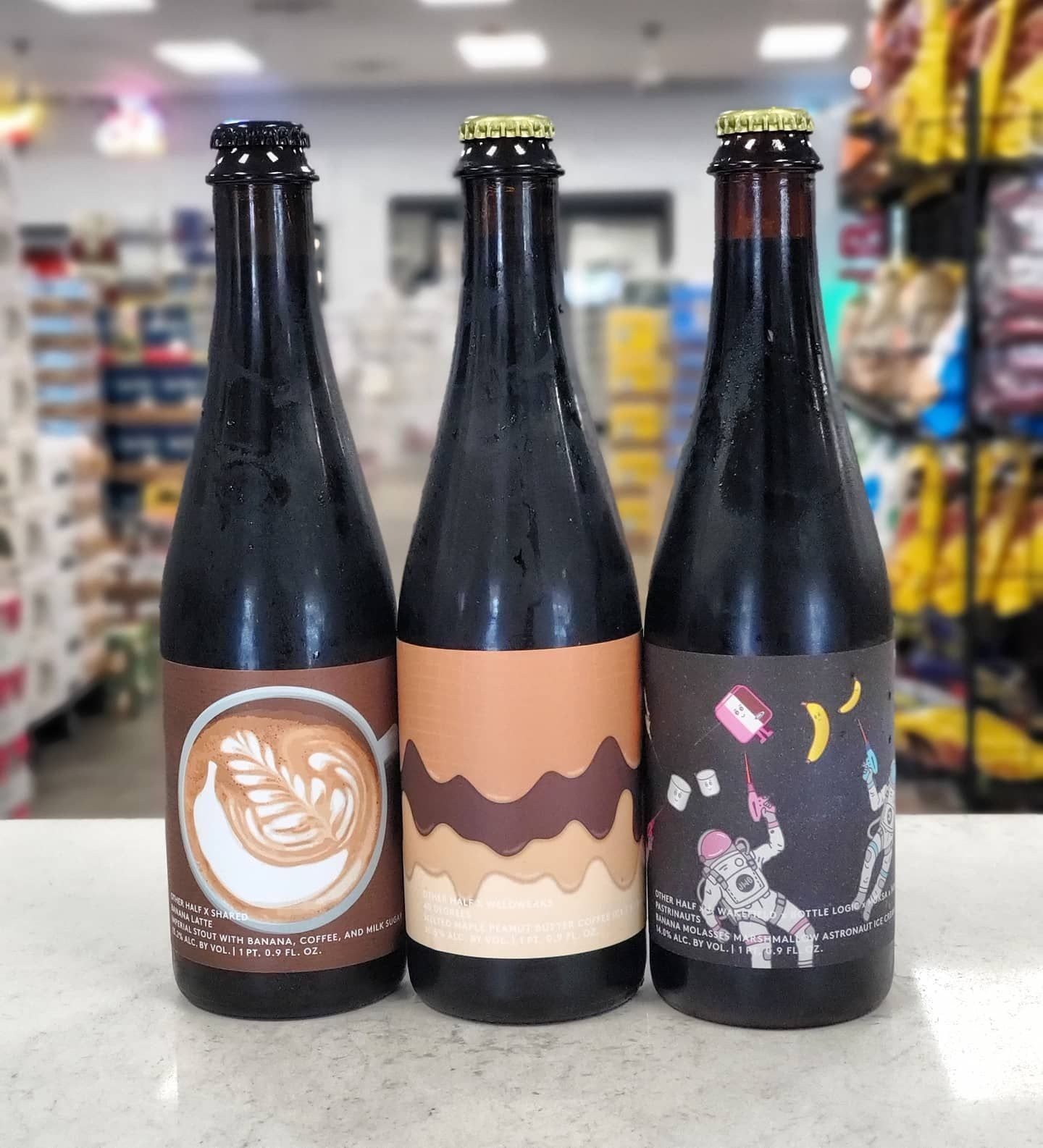 Three dark beer bottles with colorful labels in a store, featuring coffee art, layered design, and space-themed illustration.