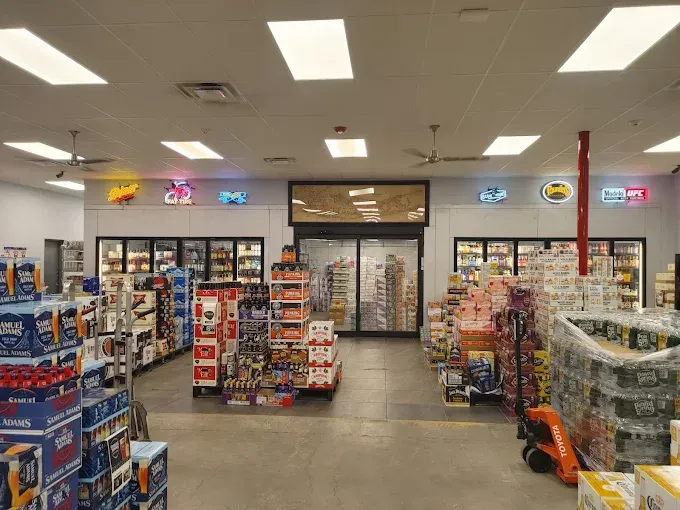 Inside a convenience store, filled with aisles of products and refrigerators. Overhead fluorescent lights illuminate the space.