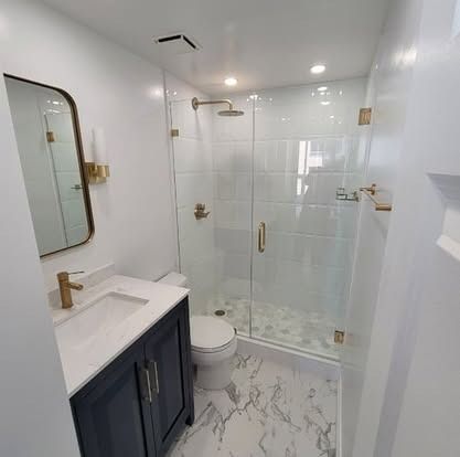 Bright modern bathroom with white tile, glass shower, vanity sink, toilet, and gold fixtures