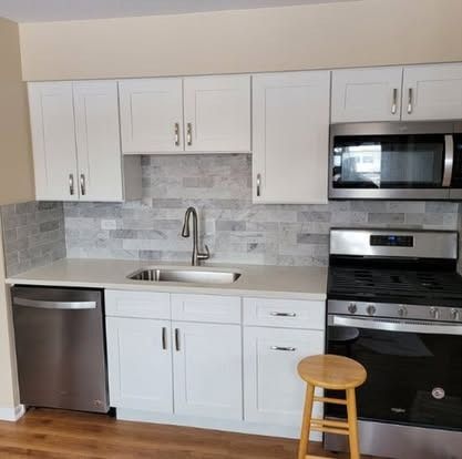 Modern white kitchen with stainless steel appliances, gray tile backsplash, and a small wooden stool.
