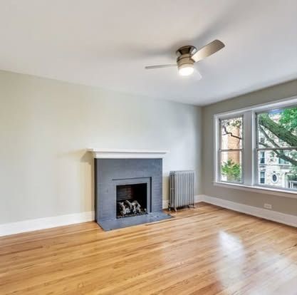 Bright empty living room with hardwood floors, gray fireplace, ceiling fan, and large window.