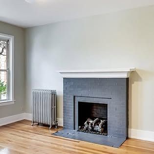 Empty living room with gray fireplace, radiator, hardwood floor, and window.