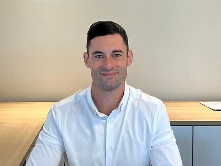 A man in a white shirt is sitting at a desk and smiling.