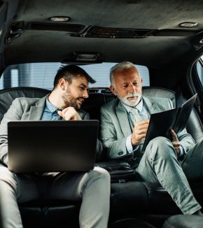 Two men are sitting in the back seat of a car looking at a laptop.
