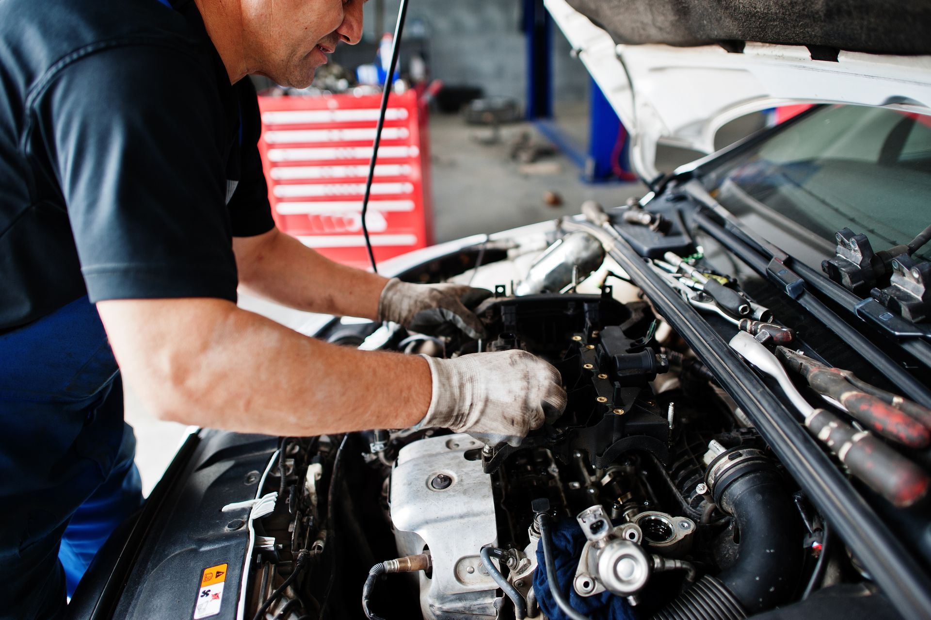 Mechanic working under the hood of a car in a garage, inspecting the engine with gloved hands