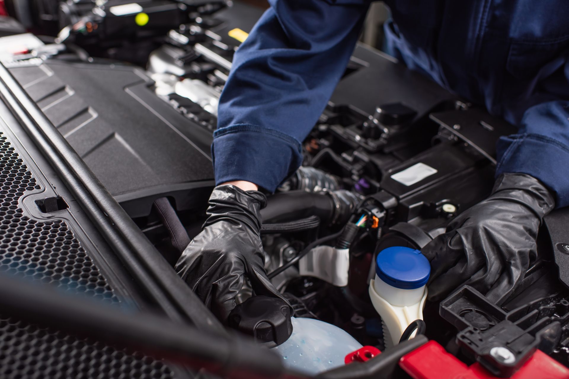 Mechanic in black gloves working under a car hood, checking fluid levels near a blue cap.