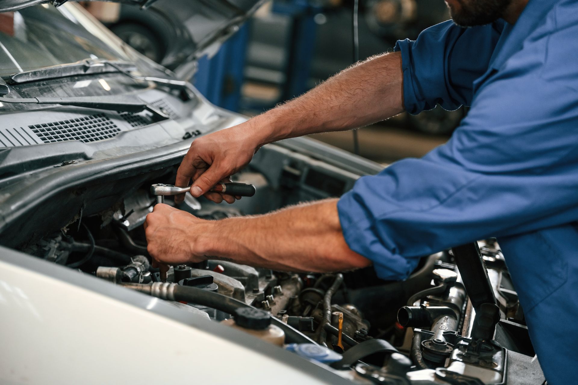 Mechanic working under an open car hood in a garage, adjusting engine components.
