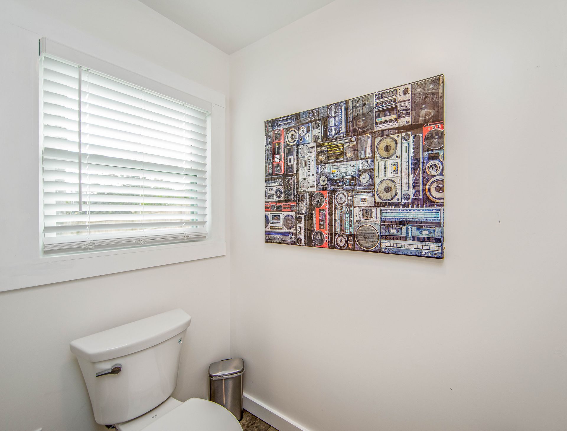 A bathroom with a toilet , trash can , and a painting on the wall.