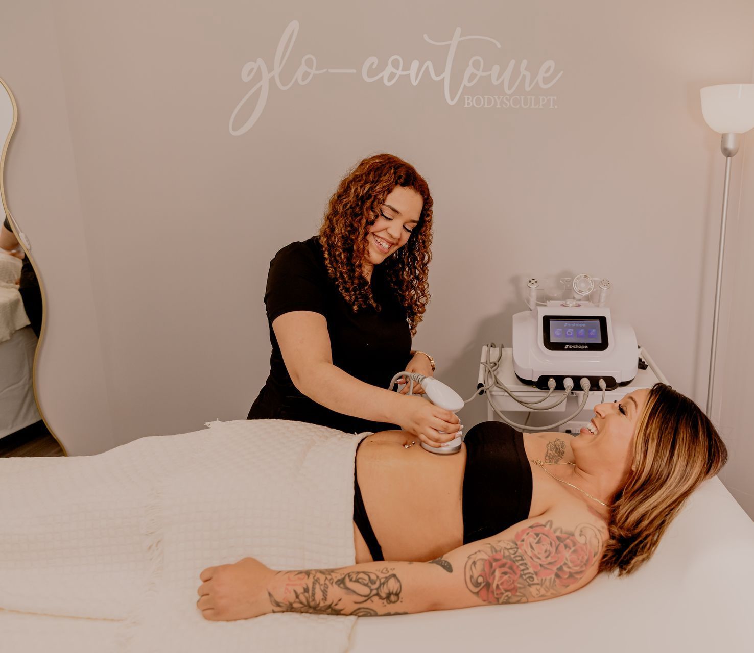 Woman receiving body contouring treatment in a spa. Practitioner uses hand-held device. Both smile.